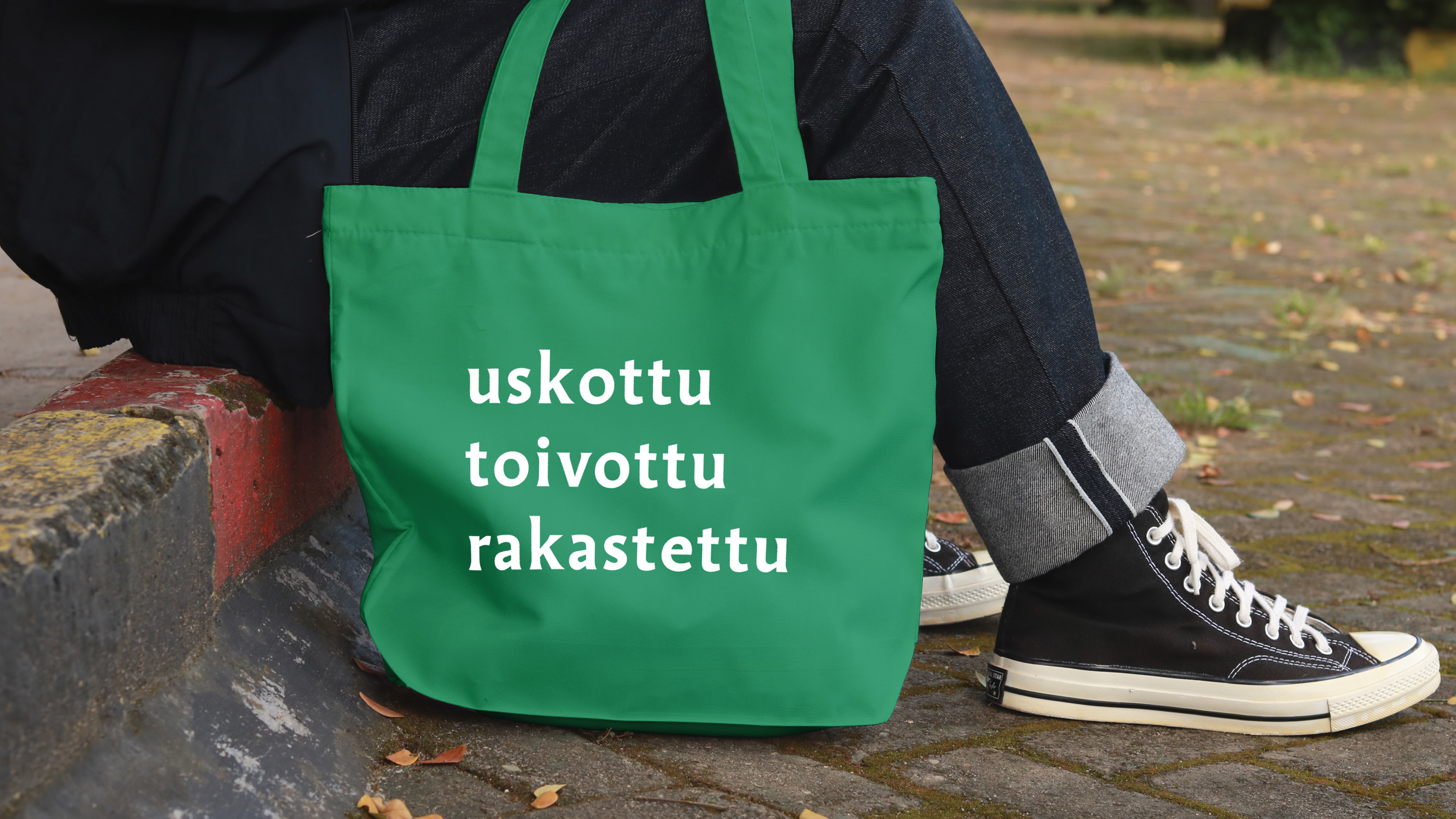 Portaalla istuva ihminen, jolla vihreä kassi vieressä. Kassissa lukee uskottu, toivottu, rakastettu